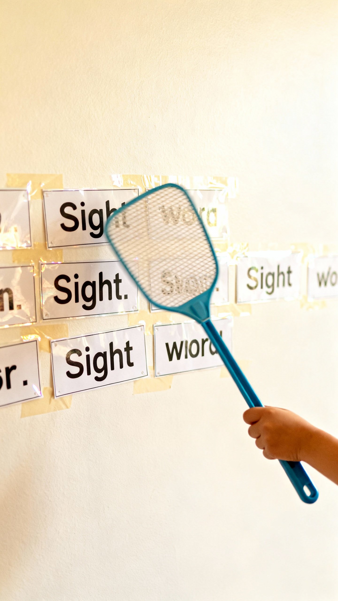 Fly swatter mid-swing toward taped sight word cards on bright wall, child’s small hand, laminated 
